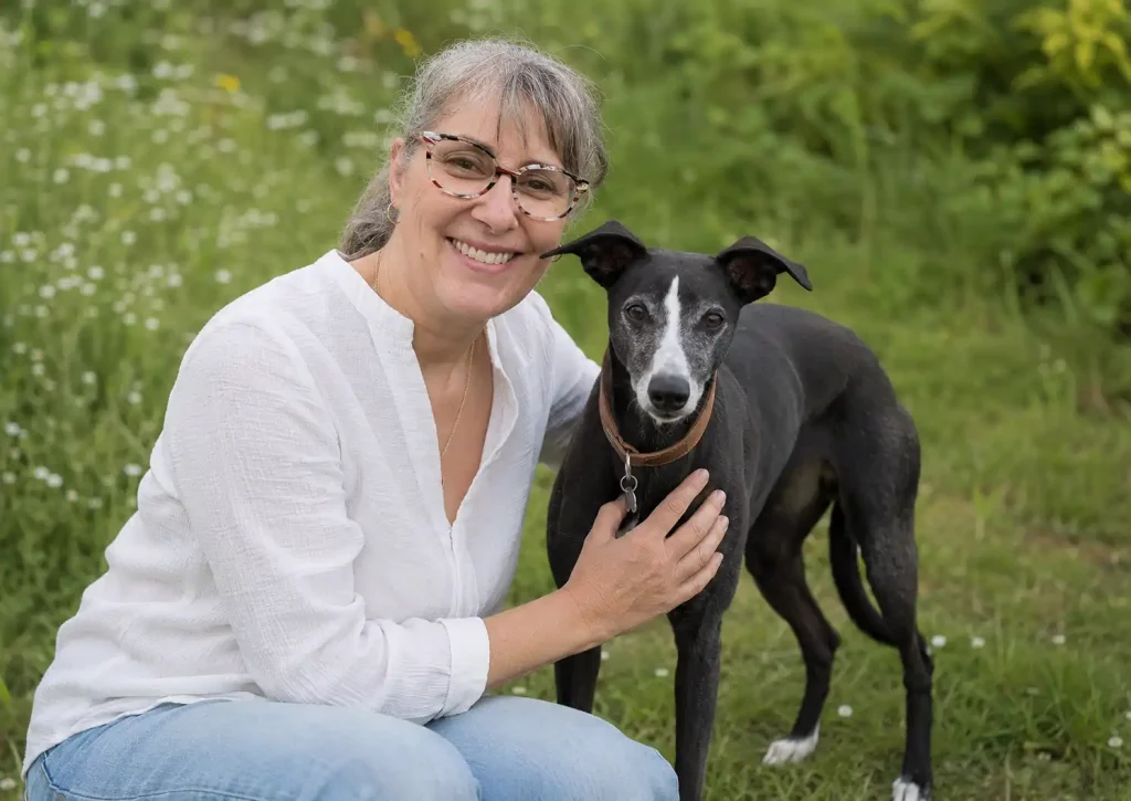 Semaine Nationale du Chien : Laura accroupie dans l’herbe avec Pimprenelle, sa Whippet noir et blanc.